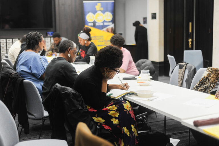 A group of people of various ages, genders, and races sit around a long table working on a creative project.