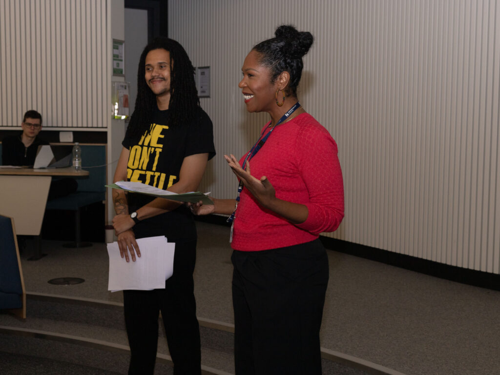 Two young people of colour stand in front of an audience, preparing to present.