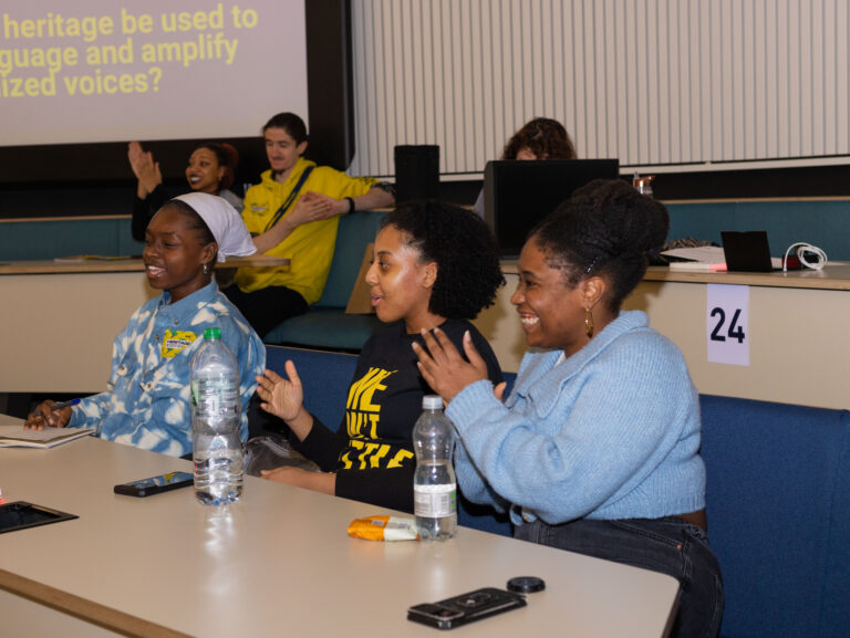 Young People of Colour sit behind desks laughing and applauding at a speaker off camera.