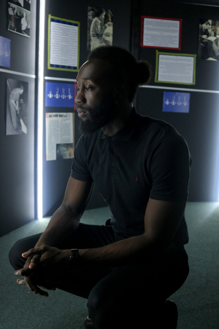 A pensive young black man crouches in an exhibition about heritage, considering the posters and pictures surrounding him.